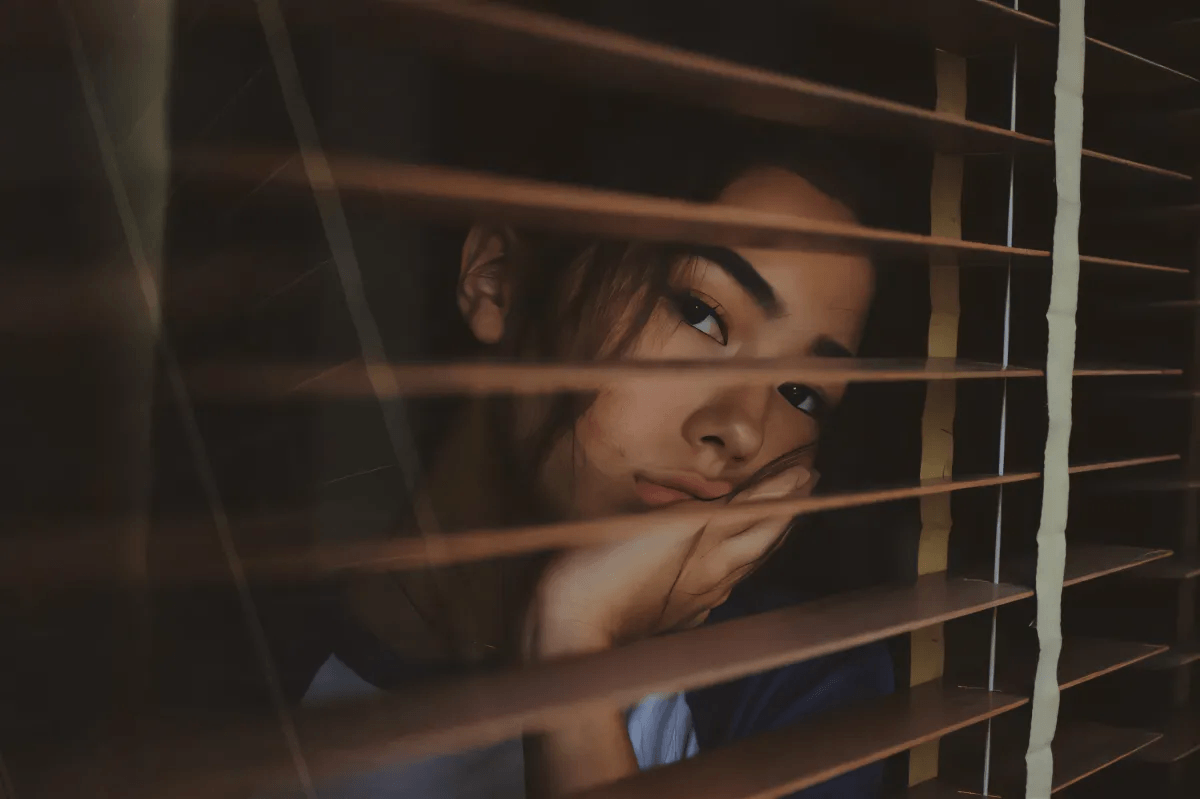 woman looking out a window with blinds
