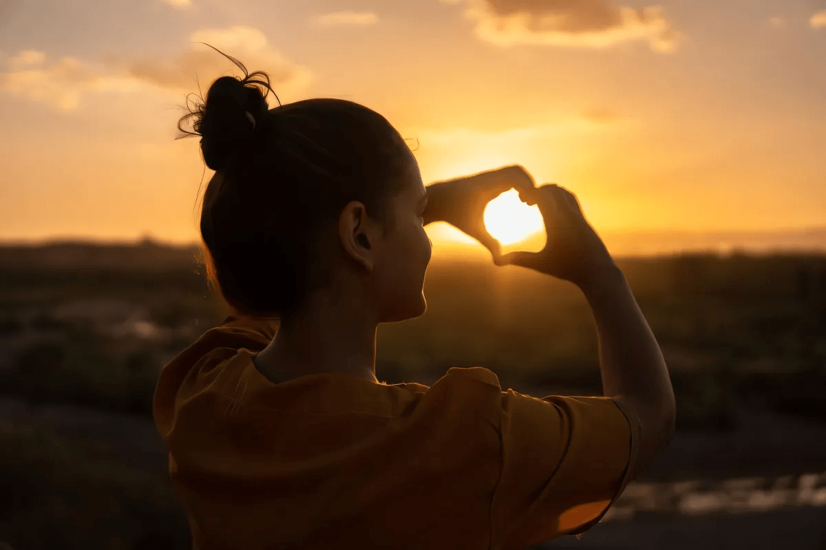 shadow of women holding heart hands over a sunset